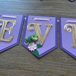 May include: A purple banner with gold glitter letters spelling "E V E". Each pennant has a gold glitter letter, with a green chameleon and pink flower embellishment. The banner is strung with a pink ribbon.