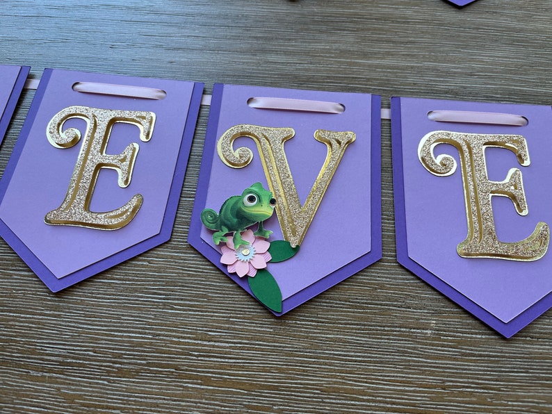 May include: A purple banner with gold glitter letters spelling "E V E". Each pennant has a gold glitter letter, with a green chameleon and pink flower embellishment. The banner is strung with a pink ribbon.