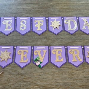 May include: A purple banner with gold glitter letters spelling "BEST DAY EVER". The banner has a light purple border and is decorated with sun and flower accents. Pink ribbons are attached for hanging.