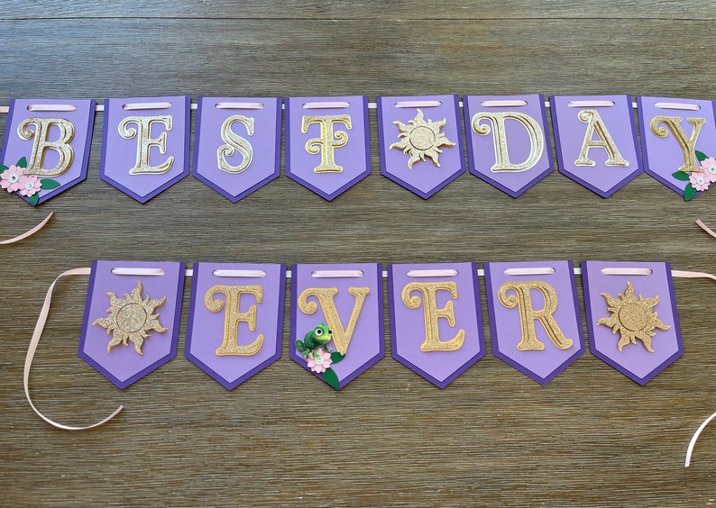 May include: A purple banner with gold glitter letters spelling "BEST DAY EVER". The banner has a light purple border and is decorated with sun and flower accents. Pink ribbons are attached for hanging.