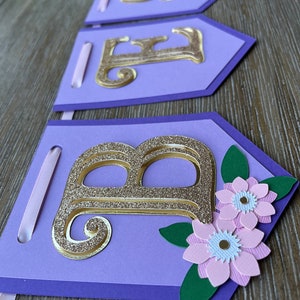 May include: A purple banner with gold glitter letters spelling "BABE". The banner has a floral design with pink flowers and green leaves. A light pink ribbon is attached to each banner piece.