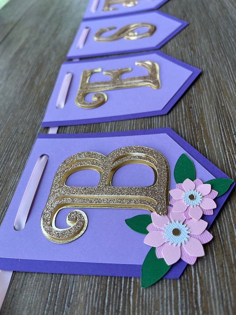 May include: A purple banner with gold glitter letters spelling "BABE". The banner has a floral design with pink flowers and green leaves. A light pink ribbon is attached to each banner piece.