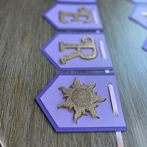 May include: A purple banner with gold glitter letters and a sun emblem. The banner is made of layered cardstock with a light pink ribbon for hanging. The letters spell out a name.