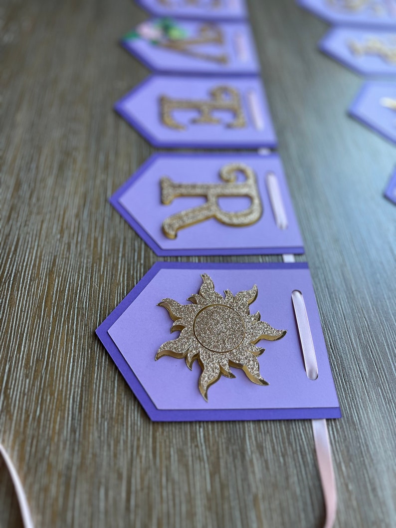May include: A purple banner with gold glitter letters and a sun emblem. The banner is made of layered cardstock with a light pink ribbon for hanging. The letters spell out a name.