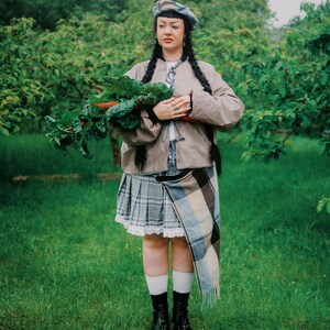 May include: A person wearing a tan jacket, plaid skirt, and black boots, holding a bundle of leafy green and red vegetables. The person is standing in a grassy area with trees in the background. They are also wearing a beret and a scarf.