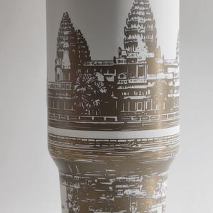 May include: White stainless steel travel mug with a gold lid and a brown silhouette of Angkor Wat temple printed on the side. The temple is reflected in the water below.