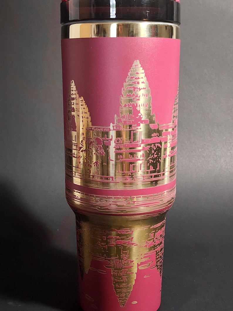 May include: A pink stainless steel travel mug with a gold-colored image of Angkor Wat, a temple complex in Cambodia.