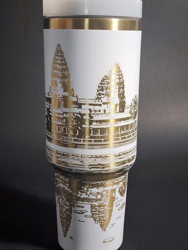 May include: White travel mug with a gold metallic design of Angkor Wat temple in Cambodia. The temple is depicted in a detailed silhouette.