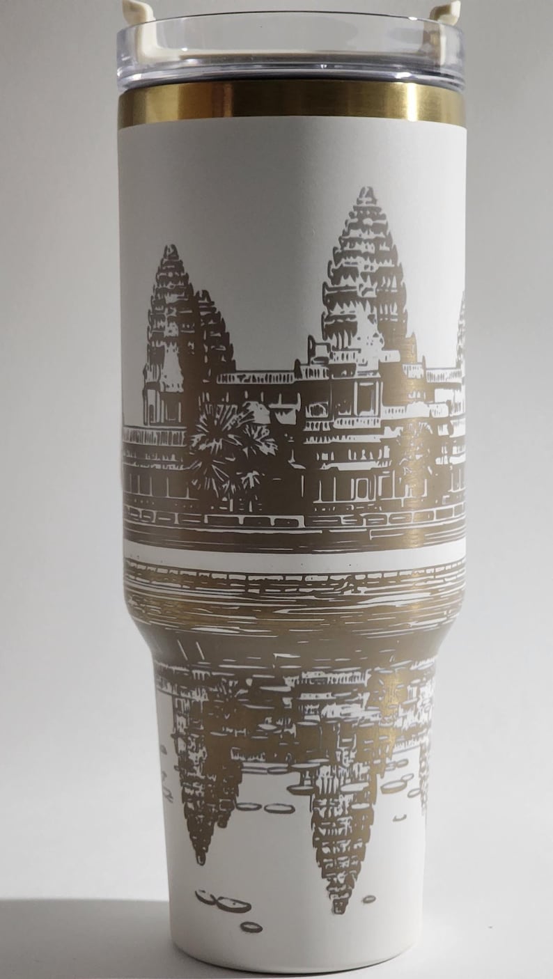 May include: White stainless steel travel mug with a gold lid and a brown silhouette of Angkor Wat temple printed on the side. The temple is reflected in the water below.