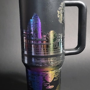 May include: A black stainless steel travel mug with a rainbow iridescent finish. The mug features an etched design of a temple with trees and a reflection in the water. The mug has a black handle.