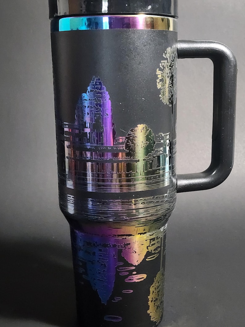 May include: A black stainless steel travel mug with a rainbow iridescent finish. The mug features an etched design of a temple with trees and a reflection in the water. The mug has a black handle.