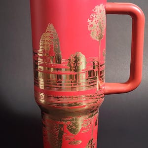 May include: Red travel mug with a gold design of Angkor Wat temple in Cambodia. The mug has a handle and a lid.
