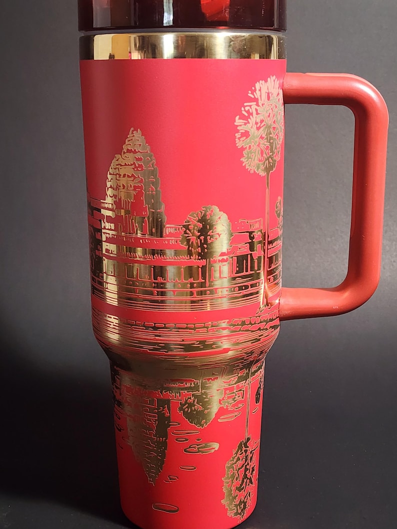 May include: Red travel mug with a gold design of Angkor Wat temple in Cambodia. The mug has a handle and a lid.