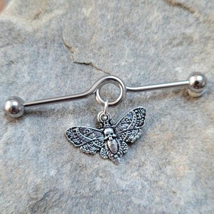 May include: Silver barbell with a dangling skull and moth charm. The barbell is curved and has two ball ends.