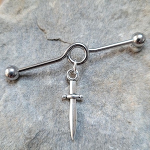 May include: Silver barbell with a dangling sword charm. The barbell is curved and has two ball ends. The sword charm is detailed and has a pointed tip.