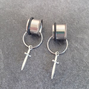 May include: Two silver ear gauges with a dangling sword charm. The gauges are circular and have a silver ring attached to the sword charm.