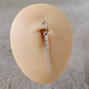 May include: Silver body jewelry with a decorative wand charm. The charm is about 2 inches long and has a detailed design.