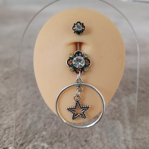 May include: A silver belly button ring with a floral design and a dangling star. The ring features a large clear gem in the center of the flower and a smaller flower at the top. The star is inside a silver hoop.