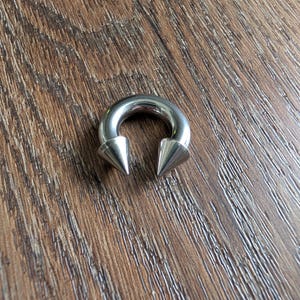 May include: A circular horseshoe-shaped body jewelry piece made of polished silver-toned metal. The ends of the circular barbell feature two pointed, cone-shaped spikes that face inward toward each other, creating a distinct and symmetrical appearance.