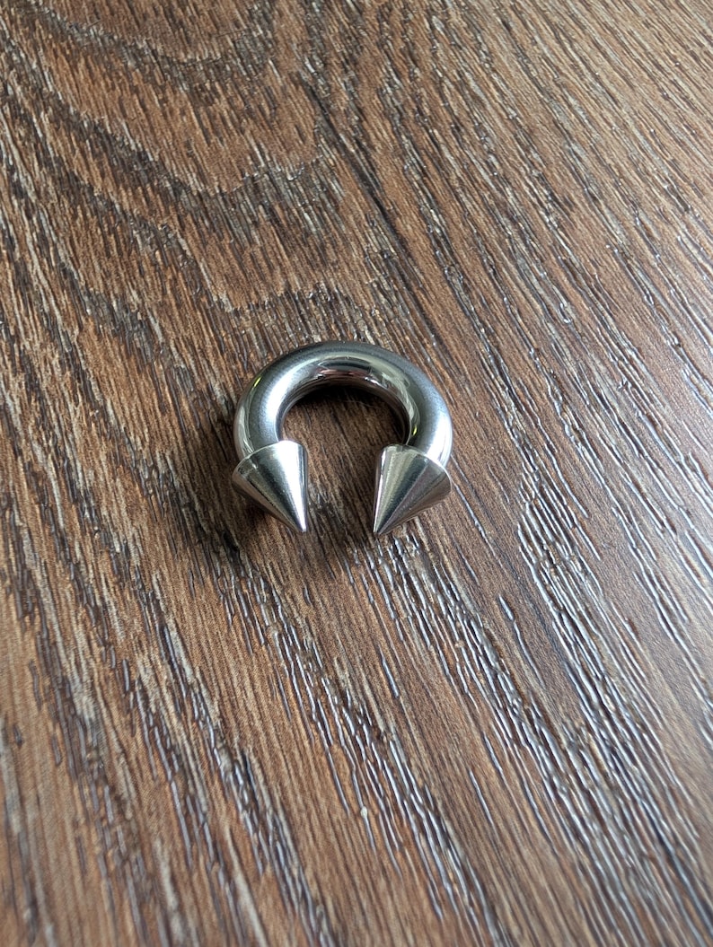 May include: A circular horseshoe-shaped body jewelry piece made of polished silver-toned metal. The ends of the circular barbell feature two pointed, cone-shaped spikes that face inward toward each other, creating a distinct and symmetrical appearance.