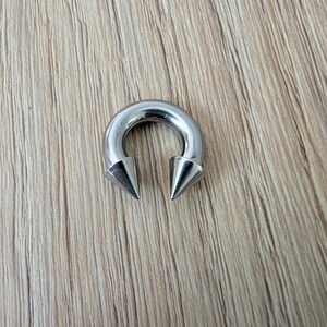May include: A circular horseshoe-shaped body jewelry piece made of polished silver-toned metal. The ends of the circular barbell feature two sharp, conical spikes that point inward toward each other, creating a distinct and symmetrical metallic design.