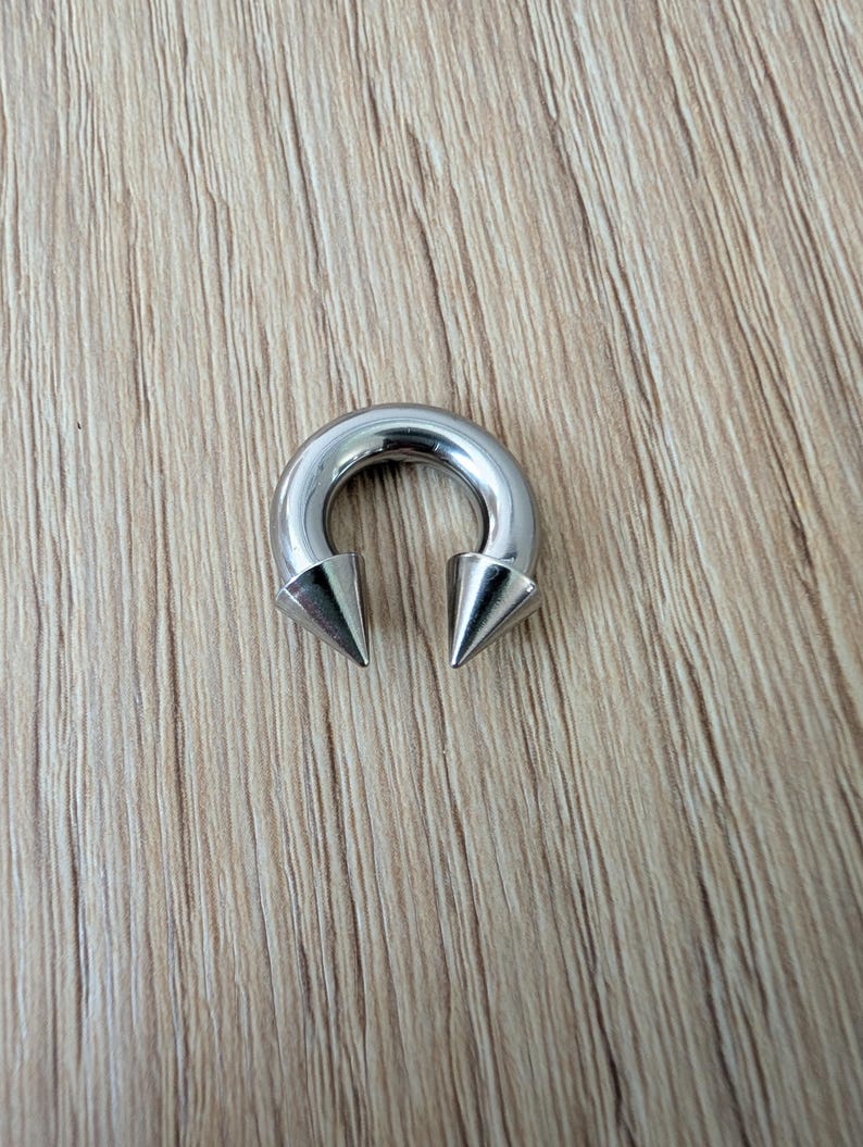 May include: A circular horseshoe-shaped body jewelry piece made of polished silver-toned metal. The ends of the circular barbell feature two sharp, conical spikes that point inward toward each other, creating a distinct and symmetrical metallic design.