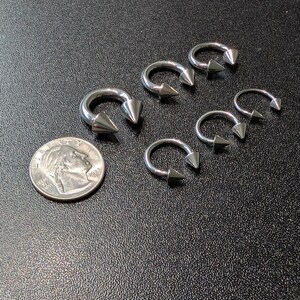 May include: A collection of five silver-toned circular barbell body jewelry pieces, each featuring two pointed cone ends. The items are arranged next to a United States dime coin for scale, showcasing their metallic finish and curved horseshoe shape.