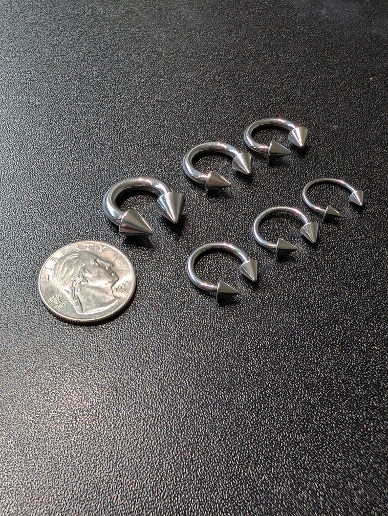 May include: A collection of five silver-toned circular barbell body jewelry pieces, each featuring two pointed cone ends. The items are arranged next to a United States dime coin for scale, showcasing their metallic finish and curved horseshoe shape.