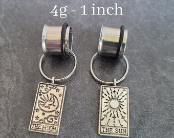May include: A pair of silver 4g - 1 inch ear gauges with a black rubber o-ring. Each gauge has a silver charm attached to it. The charms are rectangular and feature a design from the tarot deck. The charm on the left gauge features a crescent moon and the text "THE MOON". The charm on the right gauge features a sun and the text "THE SUN".