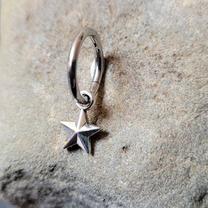 May include: Silver hoop earring with a star charm hanging from it. The earring is on a gray rock background.