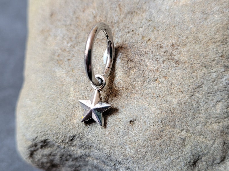 May include: Silver hoop earring with a star charm hanging from it. The earring is on a gray rock background.