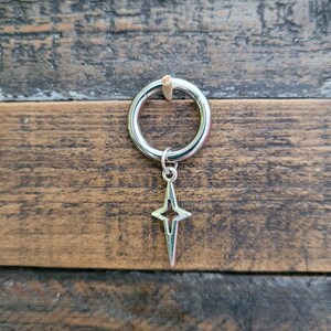 May include: A silver-colored earring with a star-shaped charm. The earring is a hoop style, and the star charm hangs from the bottom. The background is a wooden surface with a natural wood grain.