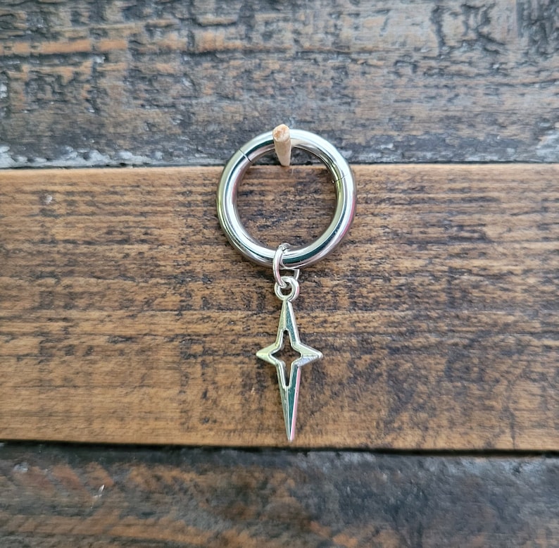 May include: A silver-colored earring with a star-shaped charm. The earring is a hoop style, and the star charm hangs from the bottom. The background is a wooden surface with a natural wood grain.