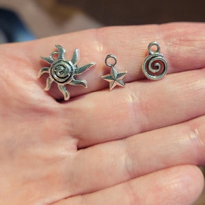 May include: Three silver charms: a sun with a spiral design, a star, and a spiral. The charms are on a hand.