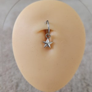 May include: A silver belly button ring with a star charm. The ring is on a light pink background.
