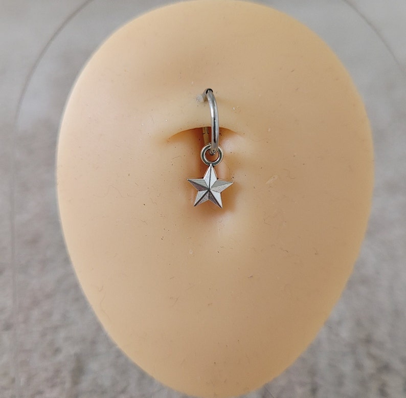 May include: A silver belly button ring with a star charm. The ring is on a light pink background.
