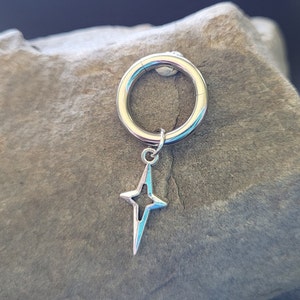 May include: A silver-colored hoop earring with a star-shaped charm. The star has a four-point design with a light blue interior. The earring is circular and has a clasp closure.