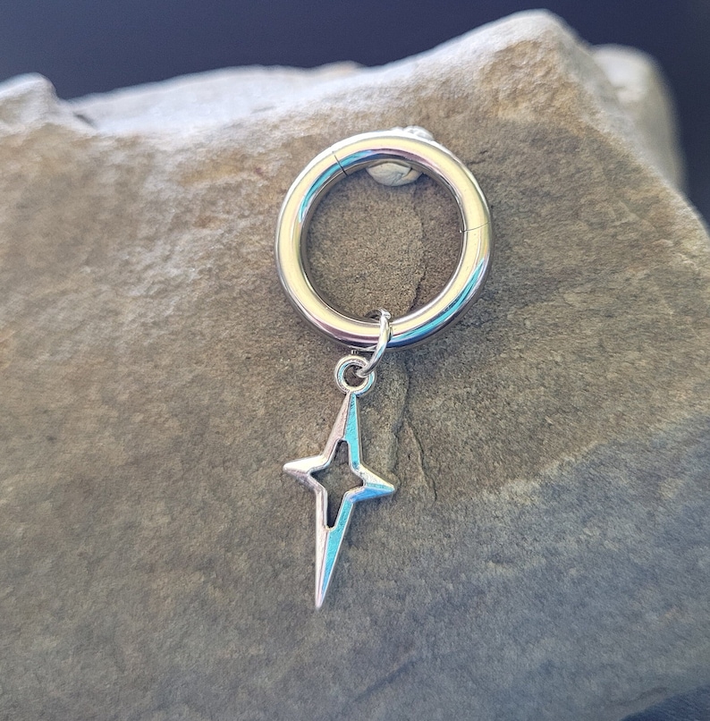 May include: A silver-colored hoop earring with a star-shaped charm. The star has a four-point design with a light blue interior. The earring is circular and has a clasp closure.