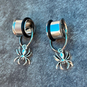 May include: Two silver ear gauges with dangling spider charms. The gauges are silver and have a shiny finish. The spiders are detailed and have a black finish.