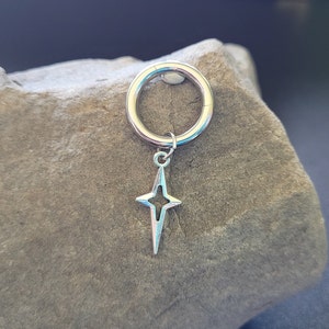 May include: A silver-colored hinged ring with a star-shaped charm. The charm has a four-pointed star design with a hollow center. The jewelry is displayed on a textured, gray stone surface.