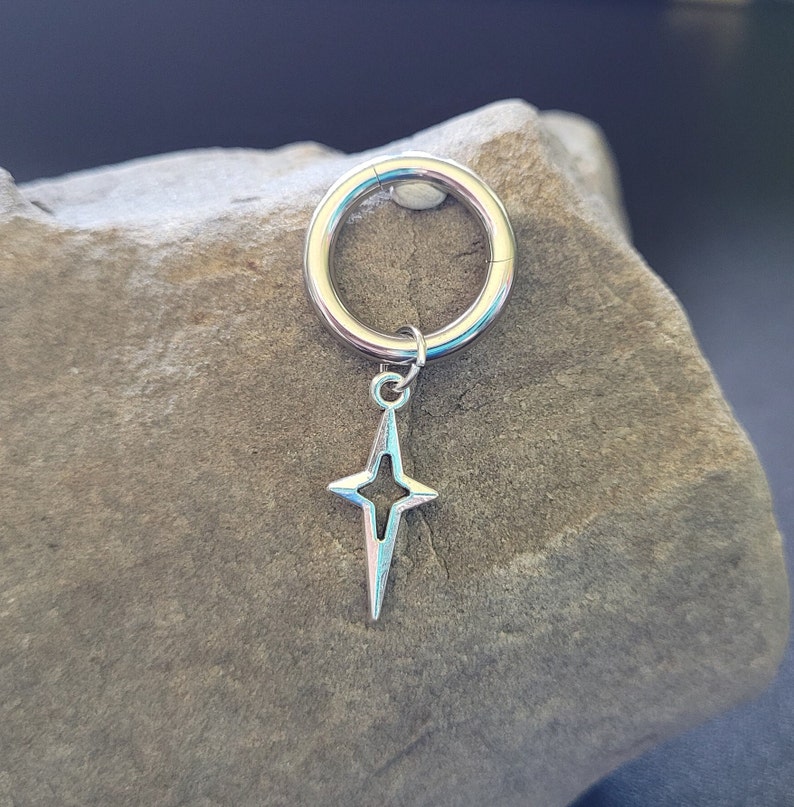 May include: A silver-colored hinged ring with a star-shaped charm. The charm has a four-pointed star design with a hollow center. The jewelry is displayed on a textured, gray stone surface.
