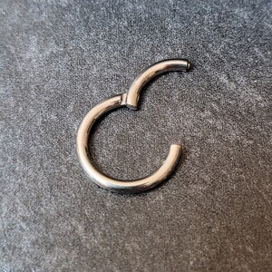 May include: A silver-colored, hinged hoop earring, open and ready to be closed. The earring is circular with a smooth, polished surface. The background is a textured, dark gray surface.