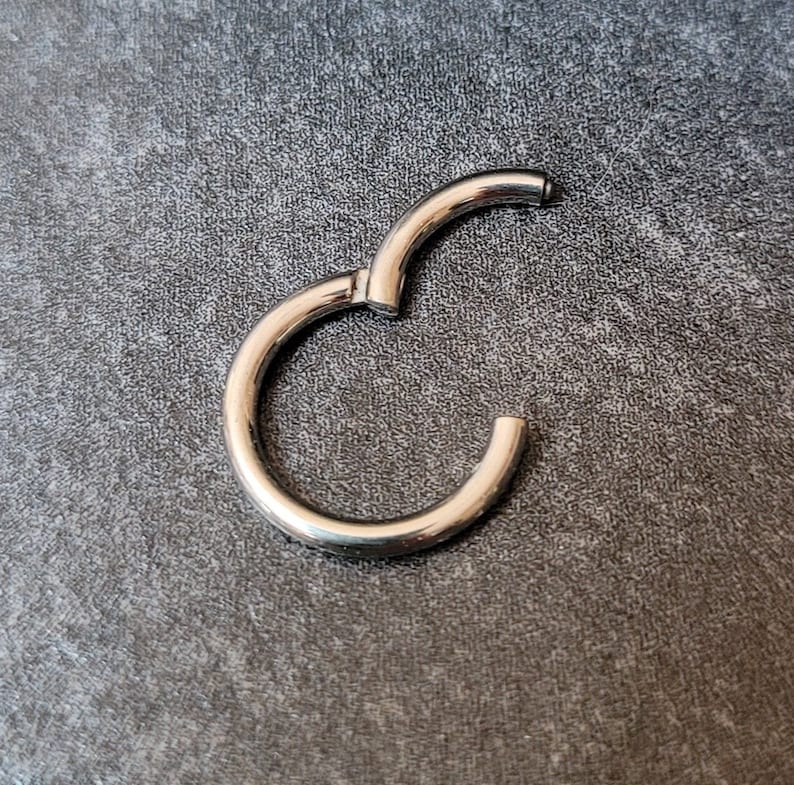 May include: A silver-colored, hinged hoop earring, open and ready to be closed. The earring is circular with a smooth, polished surface. The background is a textured, dark gray surface.