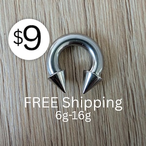 May include: A silver-colored circular barbell body jewelry piece featuring two pointed cone-shaped ends. The metal ring has a smooth, polished finish. The text 6g-16g is visible, indicating the gauge range for this specific piece of jewelry.
