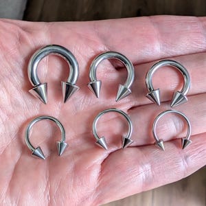 May include: Six silver-colored circular barbells arranged in two rows. Each piece of body jewelry features a smooth, curved metal hoop with a pointed, cone-shaped spike attached to each end, creating a consistent design across all six items.