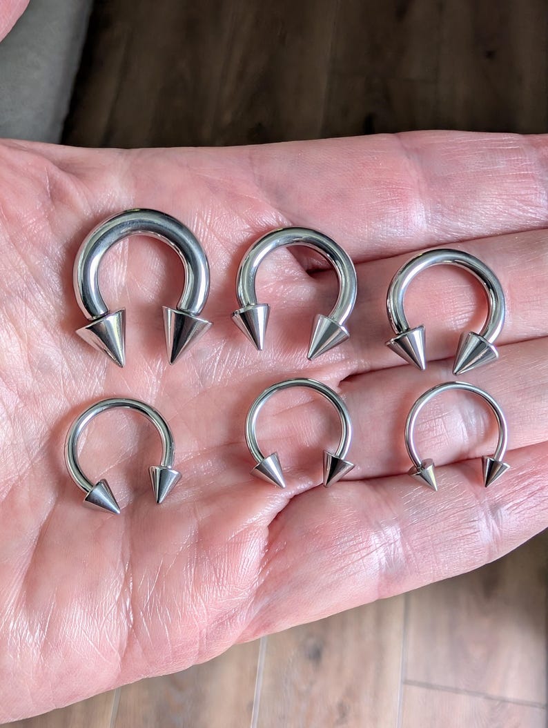 May include: Six silver-colored circular barbells arranged in two rows. Each piece of body jewelry features a smooth, curved metal hoop with a pointed, cone-shaped spike attached to each end, creating a consistent design across all six items.