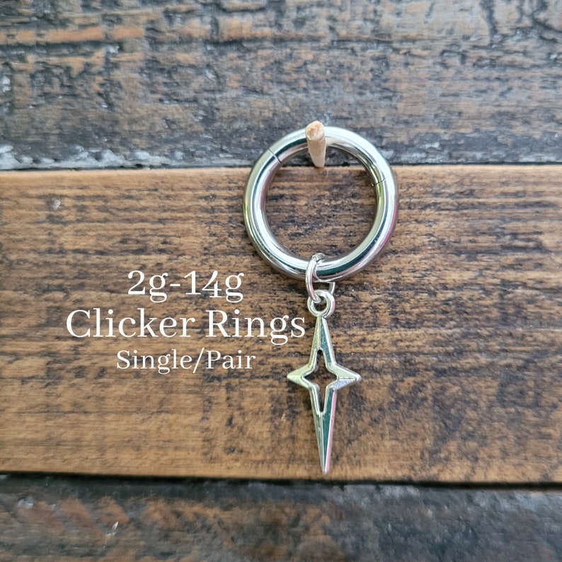 May include: A silver clicker ring with a star-shaped charm. The ring is shown against a wooden background. The text on the image reads "2g-14g Clicker Rings Single/Pair."