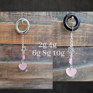 May include: Two nipple rings with a dangling charm. The charm is a pink heart with a silver star and pink beads above it. The rings are available in sizes 2g, 4g, 6g, 8g, and 10g.