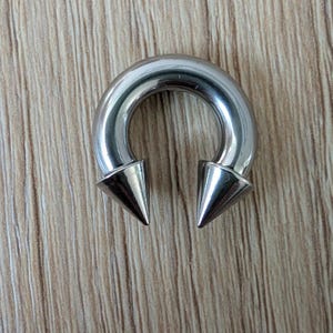May include: A circular silver-toned metal horseshoe barbell body jewelry piece featuring two pointed cone-shaped ends. The smooth, polished surface of the curved bar reflects light, and the two spikes are positioned at the bottom of the curve.
