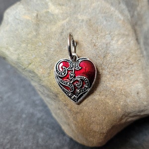 May include: A silver heart-shaped charm with a red enamel center and intricate black filigree design. The charm is hanging from a silver loop.